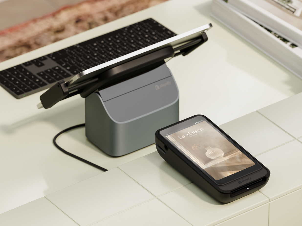 A Shopify POS Terminal sits on a white, tiled checkout counter. Behind it sits other POS accessories including a tablet, stand, and keyboard, suggesting a large retail location.